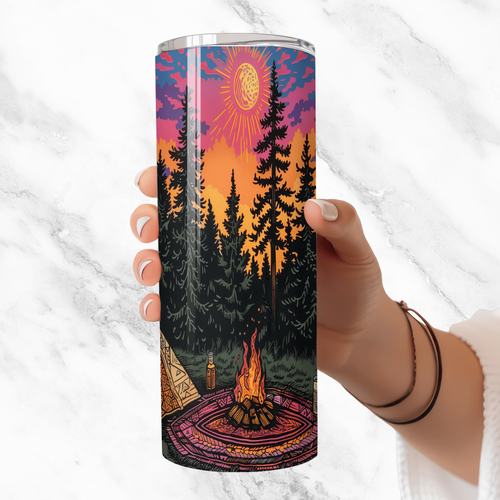 Tumbler with a forest design featuring a campfire, trees, and a sunset held by a hand against a white background.